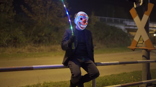 Clown with Joker Mask and glowing balloon is sitting near an railroad crossing.