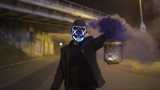 Man with purge mask and smoke grenade is holding a lantern while walking under a bridge at night.