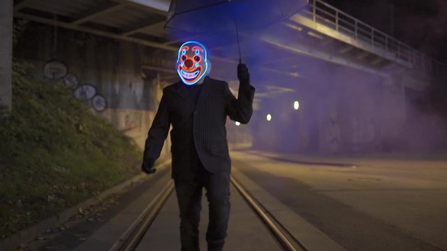 Man with clown mask and smoking umbrella is dancing under a bridge at night.