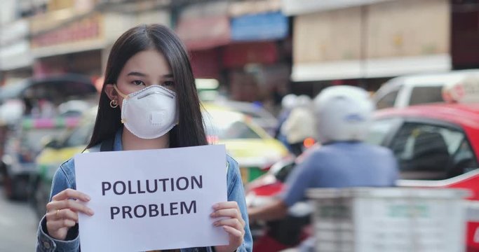 Young woman in protective mask holding pollution problem sign at city.