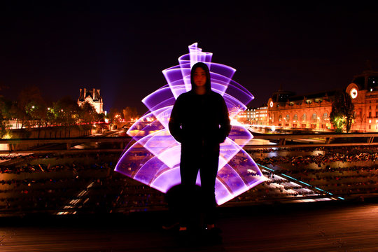Boy standing with a hood on a bridge at night. Violet light effect with a light saber in lightpainting. River Seine on background with monument. 