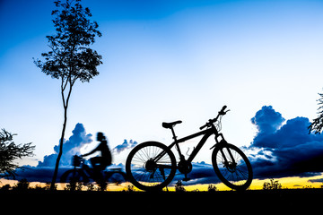 Naklejka premium Silhouette of woman is cycling through a parked bicycle in park near the lake at sunset time in summer.