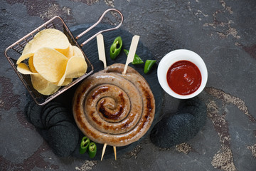 Barbecued rolled sausages on wooden skewers with potato chips, flatlay on a brown stone background, horizontal shot