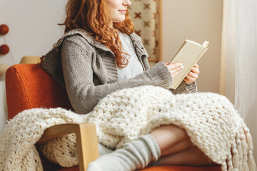 Beautiful redhead girl reading book in bed and eating cookie in cozy bedroom interior