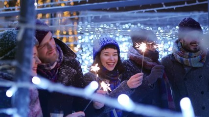 Happy friends walking with sparklers in hands and having fun on snowy winter night in festive illumination tunnel. People hanging on Christmas market. Garland lamps, carousel lights on background. - Powered by Adobe