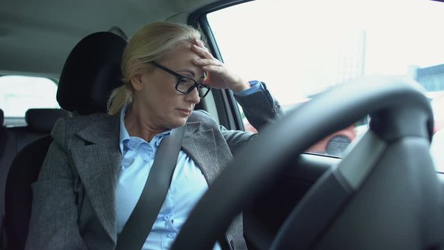 Upset aged woman in suit sitting on drivers car seat, touching head, work stress