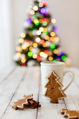 Merry Christmas! Christmas tree-shaped gingerbread cookie near cups of tea. Beautiful blurred christmas tree background with luminous garland.