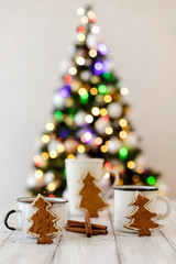 Merry Christmas! Christmas tree-shaped gingerbread cookie near cups of tea. Beautiful blurred christmas tree background with luminous garland.