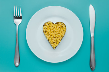 Pasta on a white plate with fork and knife on turquoise background. Heap of uncooked macaroni. Shape of heart, symbol of love. Creative romantic concept.
