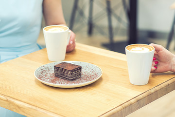 A piece of tasty chocolate cake on the table on background of hands with cups of coffee in cafe. A slice of cake on the plate at cafe.