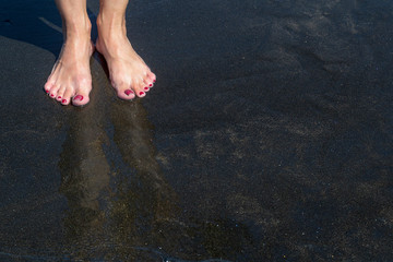 Woman's feet on a black sandy beach and reflection in the water from above.