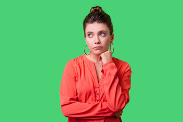 Portrait of resentful elegant woman with bun hairstyle, big earrings and in red blouse looking aside, holding her chin and pondering sadly, depressed. indoor studio shot isolated on green background