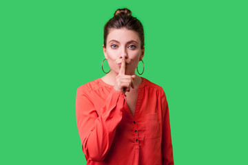 Keep silence! Portrait of elegant woman with bun hairstyle, big earrings and in red blouse making quiet gesture with finger on lips, asking for secrecy. indoor studio shot isolated on green background