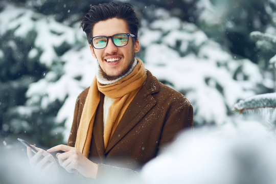 Young man in a coat on the street with a phone. Smiling young man having fun outdoors. Businessman uses a phone.  Man enjoy a winter. Dressed in a coat, sweater and scarf. Winter concept. Snowfall.