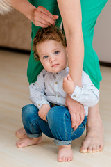 Naklejka premium a small cute child with curly blond hair sits on the floor under the feet of an adult. portrait of a little boy next to his mother