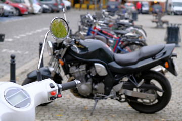 White urban scooter and group of powerful black motorcycles are parked in a row on a cobblestone pavement in the tourist center of the city.