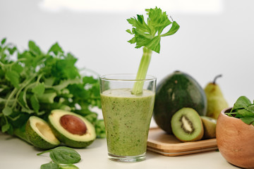 Green smoothie with a stalk of celery in a glass cup. On the table is an avocado.