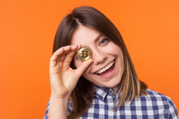 Closeup portrait of playful brunette woman with charming smile wearing checkered shirt standing covering one eye with golden bitcoin, cryptocurrency. indoor studio shot isolated on orange background