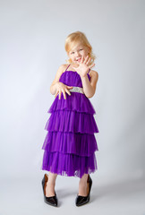 Little princess girl in fashion purple dress wearing big mothers patent-leather heels shoes in studio.Blonde girl showing and pointing up with fingers number five while smiling confident