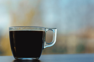 Small transparent cup of black coffee. Placed on dark wooden table in front of window. Blurred autumn sky and yellow trees in background.