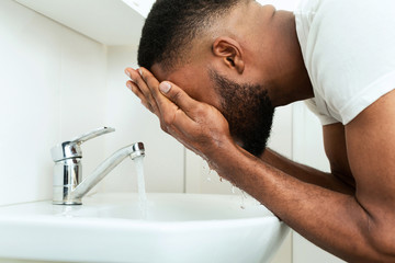 Black man washing his face, splashing water at sink in bathroom