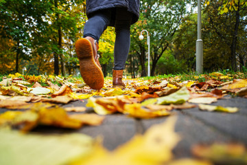 legs of a woman in black pants and brown boots walking in a park along the sidewalk strewn with fallen leaves. The concept of turnover seasons. Weather background