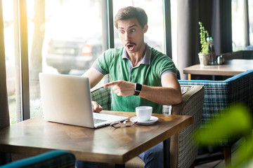 Young shocked businessman in green t-shirt sitting, working, looking and pointing at laptop display with amazed unbelievable face. business and freelancing concept. indoor shot near window at daytime.