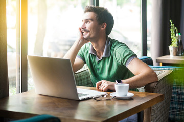 Young smiley happy businessman in green t-shirt sitting, working on laptop and looking outside, dreaming or pondering. business and freelancing dream concept. indoor shot near big window at daytime.