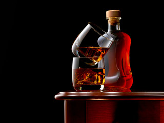 Two glasses of whiskey and ice on the wooden table. The glasses are stacked on top of each other. In the background, a closed bottle of whiskey. Low key.