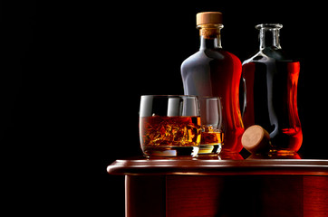 Two glasses of double whiskey and ice on a wooden table. In the background, two bottles of whiskey, closed and open. Low key.