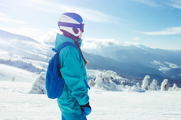 Fototapeta na wymiar A young girl enjoys beauty of winter landscape. A beautiful girl in winter clothes, a blue helmet and jacket is having a great time in the mountains. Concept of travel, leisure, freedom, sport, nature