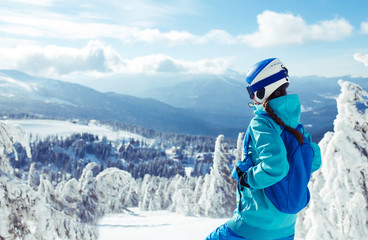 A young girl enjoys beauty of winter landscape. A beautiful girl in winter clothes, a blue helmet and jacket is having a great time in the mountains. Concept of travel, leisure, freedom, sport, nature
