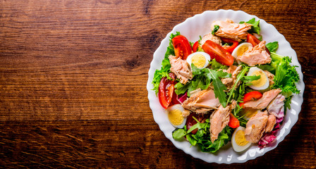 Fresh salad with fish, arugula, eggs,red pepper, lettuce, fresh sald leaves and tomato on a white plate on wooden table background