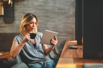 Young beautiful woman relaxing in coffee shop reading electronic book via internet on digital tablet and drink coffee - obrazy, fototapety, plakaty