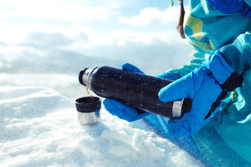 Beautiful girl in winter clothes drinking tea on the background of snow-capped mountains on the sunny day. She is wearing a blue helmet and a jacket. Concept of travel, mountains, leisure.