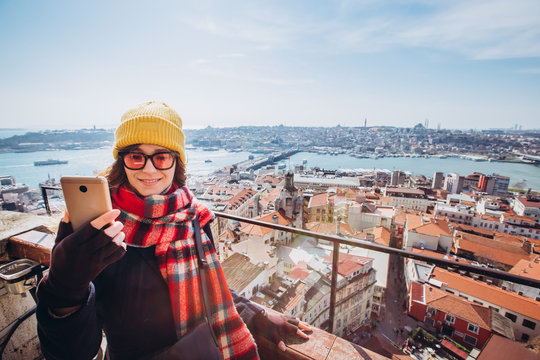 Young smiling girl on the background of Istanbul panorama on a sunny spring day. A girl in yellow hats takes a selfie on the phone in Istanbul from the observation deck of the Galata Tower.