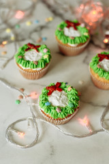 Christmas cupcakes with green Christmas wreath of cream on marble background with garland. Christmas sweets decorated with mastic