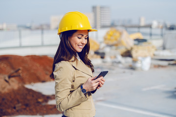 Gorgeous caucasian female architect in beige jacket and with protective helmet on standing at construction site and using smart phone.