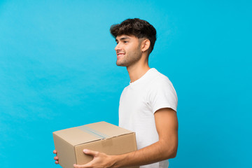 Young handsome man over isolated blue background holding a box to move it to another site