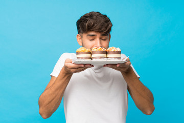 Young handsome man over isolated blue background holding mini cakes enjoying the smell of them