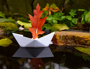 A white paper boat with a red sail on the dark water in a small pond.