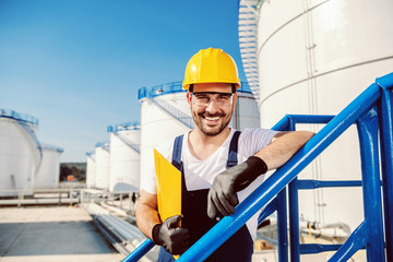 Smiling handsome caucasian worker in overall and with helmet on head leaning on the railing and holding folder with documents. Oil production.