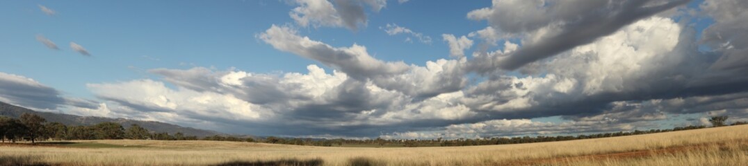 Fototapeta premium Panoramic view of large open dry drought affected farm fields under stretching cloud filled blue skies over properties in rural New South Wales, Australia