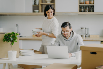 Young brunette woman unpacks boxes with personal belongings, places plates in cupboard, looks attentively at laptop computer where husband works. Family couple relocate in new modern apartment