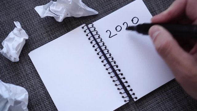A male hand writes 2020 GOALS in a notebook on a gray textured table surface. Crumpled sheets of paper are nearby. The concept of a complex creative search for planning in the new years