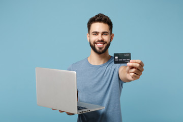 Young smiling handsome man in casual clothes posing isolated on blue wall background, studio portrait. People lifestyle concept. Mock up copy space. Hold in hands laptop pc computer, credit bank card.