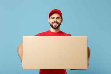 Delivery man in red uniform isolated on blue background, studio portrait. Male employee in cap t-shirt print working as courier dealer hold empty cardboard box. Service concept. Mock up copy space.