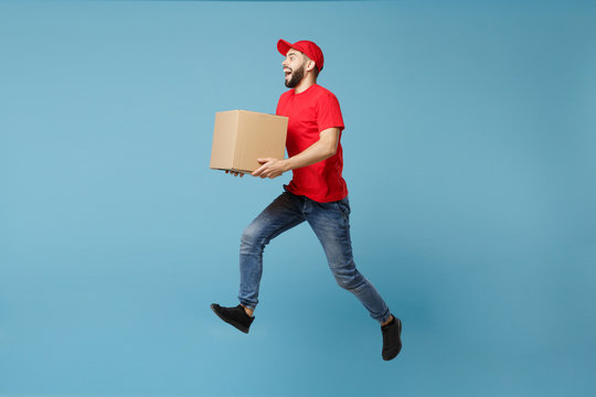 Delivery man in red uniform isolated on blue background, studio portrait. Male employee in cap t-shirt print working as courier dealer hold empty cardboard box. Service concept. Mock up copy space.