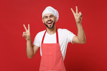 Cheerful young bearded male chef cook or baker man in striped apron white t-shirt toque chefs hat posing isolated on red background. Cooking food concept. Mock up copy space. Showing victory sign.