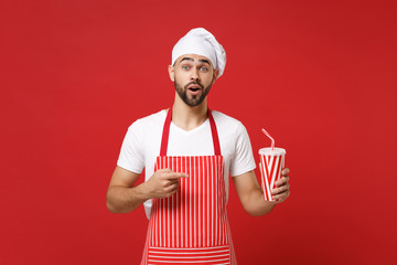 Shocked male chef cook or baker man in striped apron white t-shirt toque chefs hat isolated on red background. Cooking food concept. Mock up copy space. Pointing index finger on cup of cola or soda.